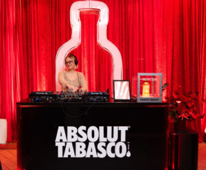 DJExperience The Fusion: ABSOLUT TABASCO Arrives In Canada! [Photos] wearing headphones mixes music at a red Absolut Tabasco event, standing behind a black mixer with a large Absolut Tabasco sign
