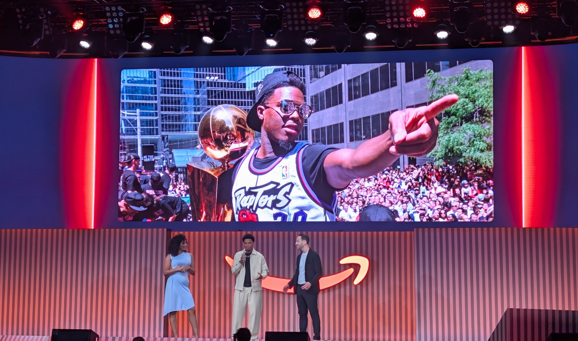 Stage with three presenters in front of a striped wall; a large screen behind shows a man in a Raptors jersey pointing toward the crowd, wearing sunglasses and a backward cap.