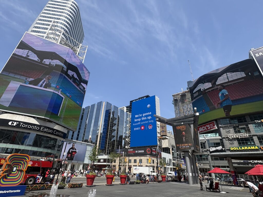 View of Yonge-Dundas Square in Toronto with large digital billboards, skyscrapers, and pedestrians in a sunny plaza.