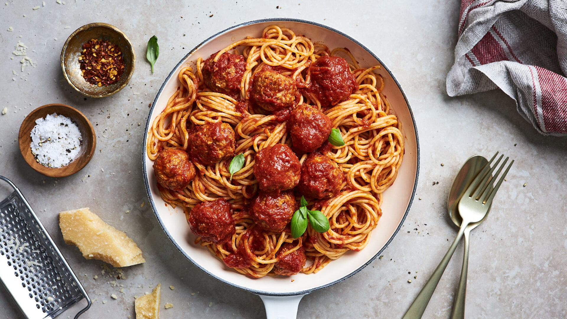 Skillet pasta with tomato sauce and meatballs garnished with basil, shown on a light counter with cheese and utensils nearby