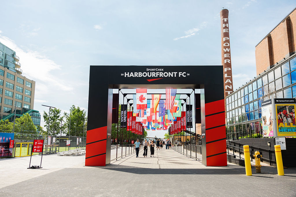 Entrance arch for HarbourFront FC with colorful international flags hanging overhead and people walking through the plaza.
