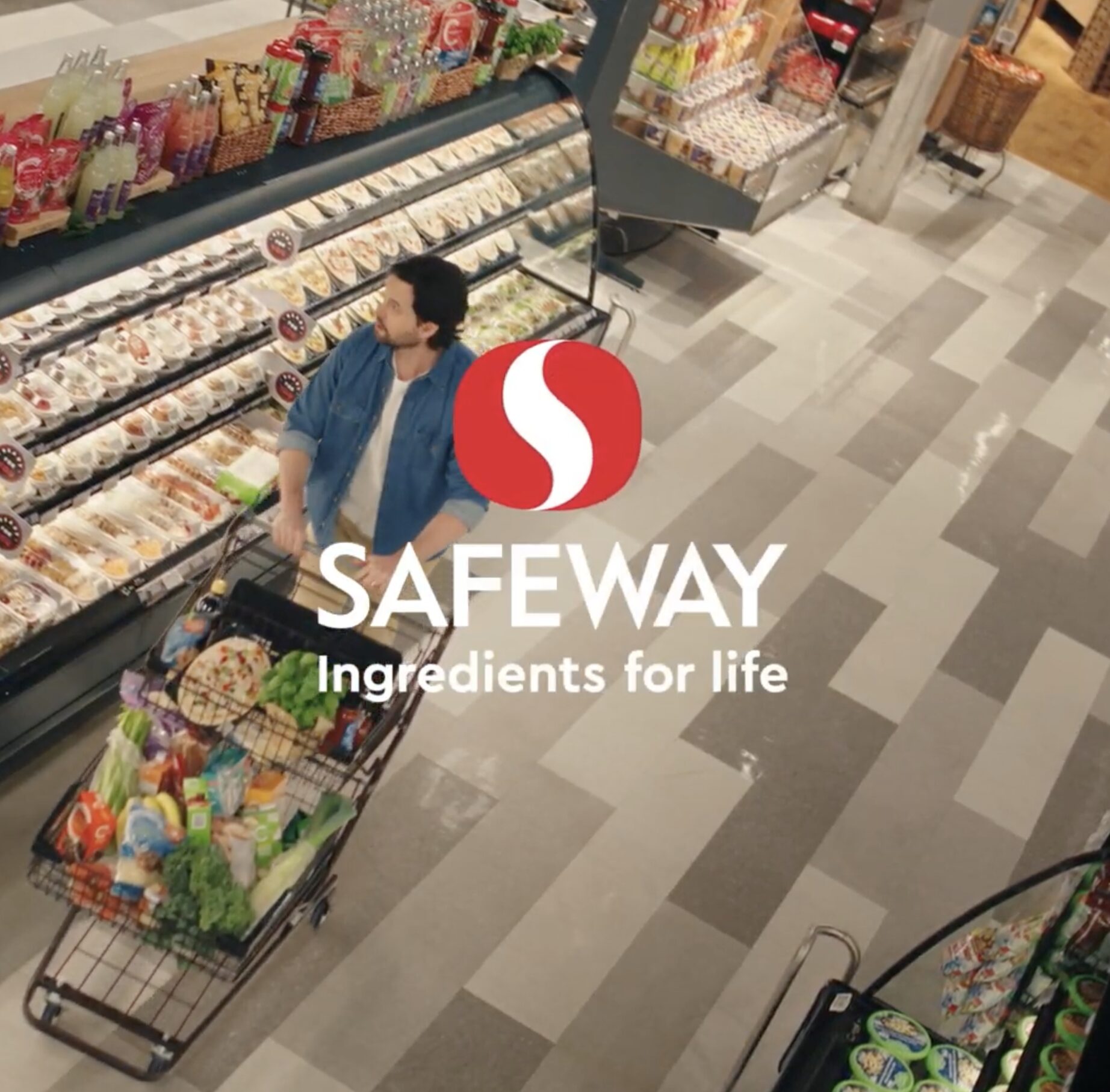 Safeway Goes All In on Humour And Relatability For New "That’s The Way" Summer Campaign - Canadian Ad Insider Man in a blue denim shirt pushes a shopping cart along a refrigerated aisle in a Safeway store.