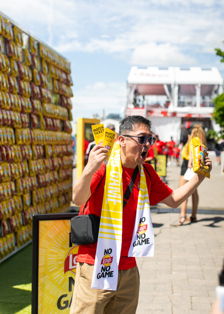 Man in a red shirt and sunglasses at an outdoor event, waving two yellow Lay’s match-ticket cards while holding a bag of chips.