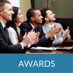 Diverse professionals applauding at an awards ceremony, seated at a table, with a blue 'AWARDS' banner beneath them.