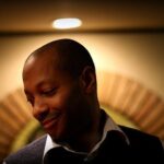Man with a gentle smile looking downward in a warm, softly lit room with a blurred background behind him.