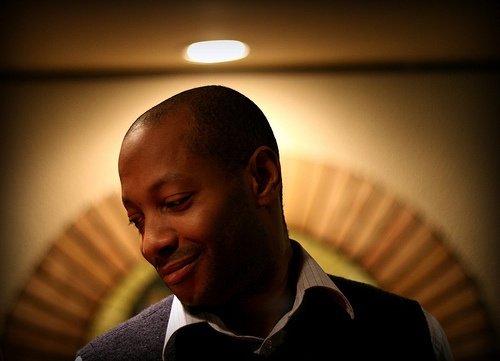 Man with a gentle smile looking downward in a warm, softly lit room with a blurred background behind him.