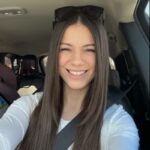 Smiling woman wearing a white top in a car, seatbelt on, sunglasses resting on her head, taking a selfie.