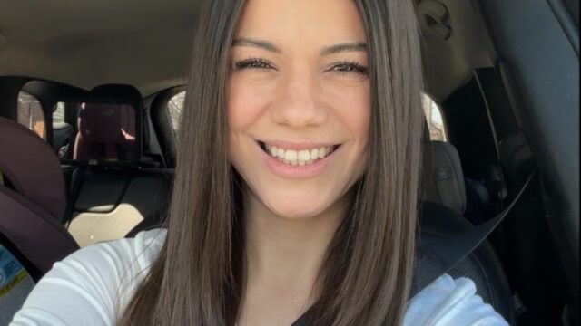 Smiling woman wearing a white top in a car, seatbelt on, sunglasses resting on her head, taking a selfie.