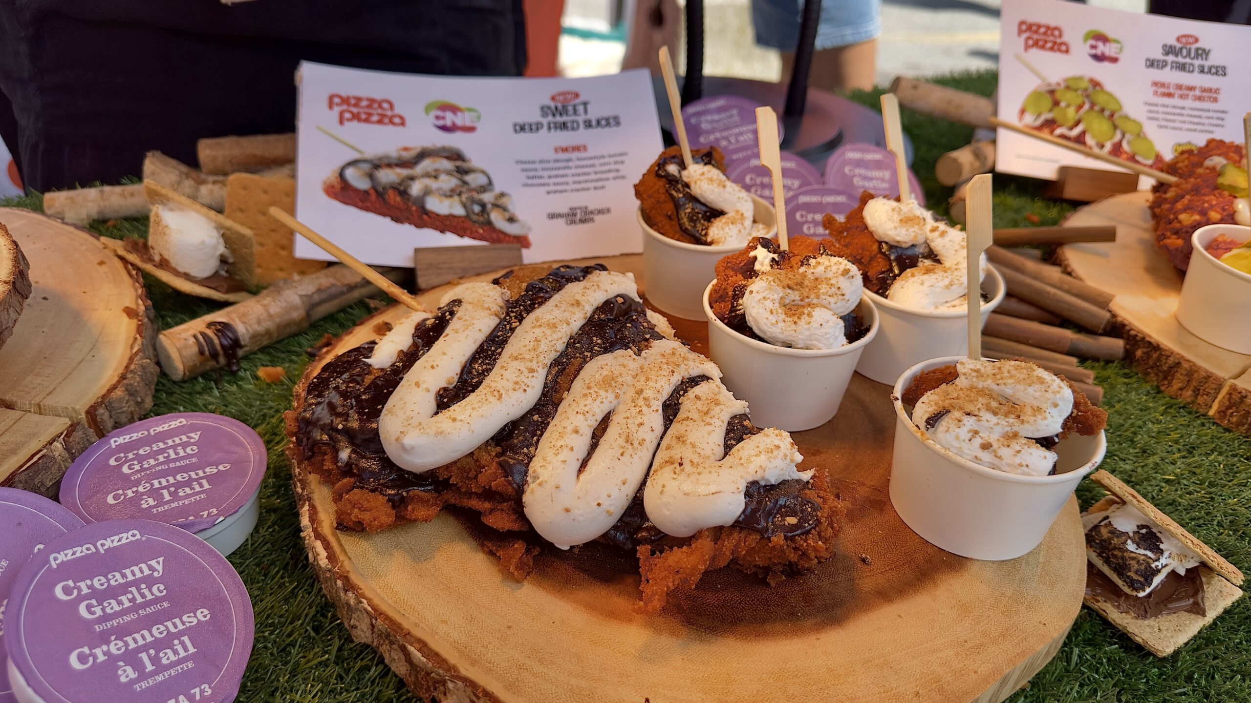 Rustic display of fried treats (churros with white icing) on a wooden board, with small cups topped with whipped cream and cinnamon, and purple garlic dipping sauce containers nearby.