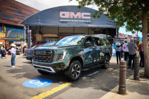 Green GMC pickup truck on display at an outdoor car show with a large GMC stage banner in the background and attendees nearby.