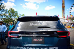 Rear view of a dark blue GMC SUV with Calgary Stampede script on the rear window and'Official Partner of the Calgary Stampede' banner on the tailgate, at a fairground.