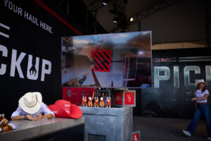 GMC promotional booth with a large TV screen, a person signing at a table surrounded by plush moose toys and red GMC gear.