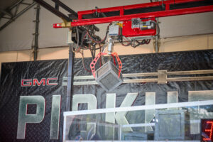 Overhead red crane and hoist machinery lifting a heavy metal block in an industrial workshop with a GMC backdrop.