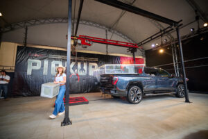 Indoor vehicle showroom with a gray GMC pickup on display, red overhead machinery, and a woman in a white hat standing near a banner.