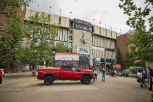 Red lifted pickup truck parked in front of GMC Stadium during an outdoor event, with banners and people nearby.