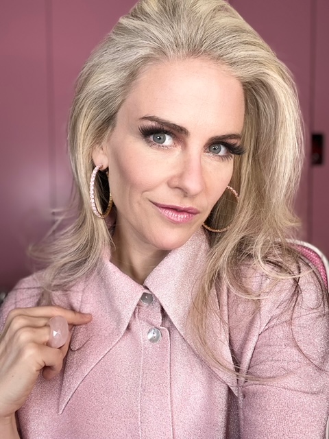 Portrait of a woman with long blonde hair, wearing a pink collared sweater and large hoop earrings, looking at the camera against a pink background.