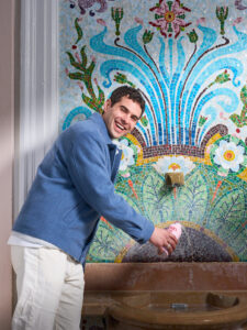 Smiling man in a blue jacket leaning toward a colorful mosaic fountain backdrop.