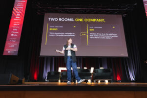 Male presenter on stage delivering a talk with a large screen displaying'TWO ROOMS. ONE COMPANY.' behind him.
