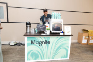 Barista standing behind a coffee kiosk with a teal Magnite banner, espresso machine, and stacked turquoise cups on the counter.
