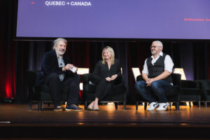 Three panelists seated on a stage in discussion, smiling, with a large screen overhead showing'QUEBEC → CANADA'.