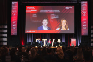 Two speakers sit on a stage in conversation, a large screen behind them displays event branding and speakers' photos. Audience watches from a dimly lit auditorium.