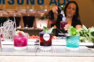 Three colorful cocktails on a metal bar counter with a bartender pouring a drink in the background.