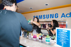 Bartender hands a green cocktail to a customer at a Samsung Ads bar, with two staff serving behind the counter.