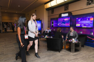 Two women walk past a Roku booth in a conference lounge with attendees seated in black chairs and large screens in the background.