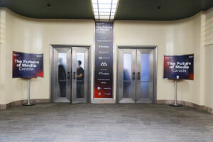 Conference lobby with two metal elevators on either side of a tall sponsor banner that reads The Future of Media Canada, plus side display screens.