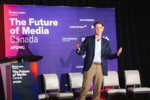 Male speaker in a navy blazer and tan pants presenting on a stage with a purple backdrop reading'The Future of Media Canada'. A podium on the left and empty chairs behind him.
