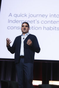 Presenter on stage in a white shirt and dark cardigan, gesturing with hands while a large screen with text looms behind him.