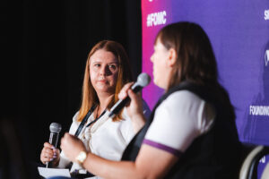 Two women on a stage in a panel discussion; one speaks into a microphone while the other listens, with a purple backdrop and visible hashtag text behind them.