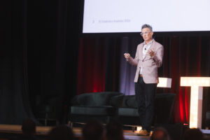 Male speaker in a blazer giving a presentation on a stage with a projector screen overhead and seating in the background, audience out of focus.