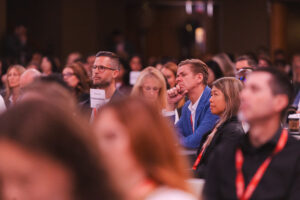 Audience at a conference; man with glasses in a blue blazer rests his chin on his hand, listening beside a woman in a black blazer.