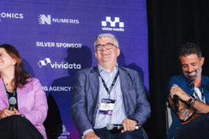 Panelists on stage at a conference; center man with glasses and a badge smiles, holding a microphone with a purple sponsor backdrop behind him.