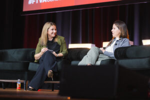 Two women sit on dark green couches on a stage, engaged in a panel discussion with a red-lit screen above them.