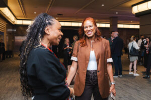 Two women converse and smile at a networking event in a conference hall, with attendees in the background