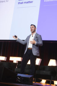 Male presenter on stage delivering a talk, gesturing with one hand and holding a remote control.