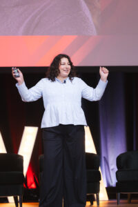 Woman presenting on stage, holding a remote in one hand and gesturing with the other, colorful stage lights behind her.