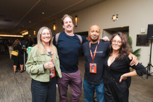 Four adults posing together at an indoor event, wearing lanyards and smiling for the camera.