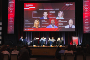 Panel discussion on a stage at a conference, with a large screen behind showing'Panel: Growing Brands in 2026' and headshots of speakers.]? Wait not.