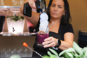 Bartender pouring a purple cocktail with dry-ice fog into a short glass, garnished with a citrus wheel.