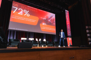 Male presenter on a conference stage beside a large screen displaying a red slide about US dominance, showing 72% and 28% data bubbles.