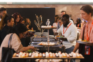 People line up at a buffet with chafing dishes and desserts during a conference event, plates in hand and conversations happening nearby.