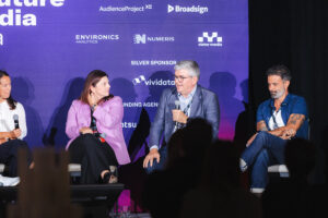 Panel discussion on a stage with four people seated, one holding a microphone, against a purple sponsor backdrop; audience silhouettes visible in the foreground.