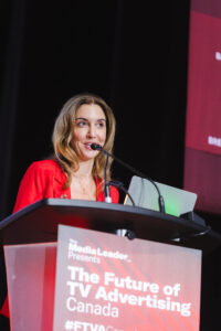 Woman in a red blazer speaks at a podium with a microphone, presenting at'The Future of TV Advertising Canada' conference.