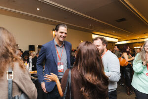 Two men and a woman converse at a conference networking event, wearing name badges in a busy room.