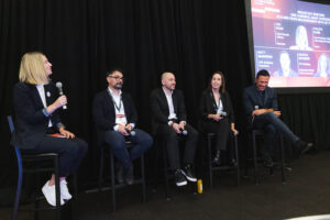 Panel of five speakers seated on a stage during a discussion, each holding a microphone, with a slide screen in the background.