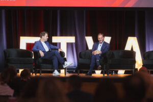 Two men in suits sit on stage in black chairs during a talk, with large illuminated letters behind them.