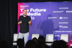 Speaker on stage delivering a talk at a media conference, with a large purple backdrop reading'The Future of Media' and empty chairs nearby.